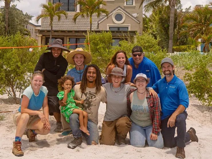 Group of eleven people, diverse ages, smiling on a sandy beach with palm trees and a house in background.