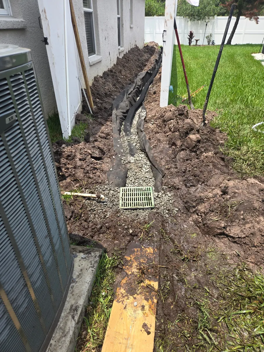 Open trench with drain pipe, filter fabric, gravel, and a catch basin grate along the side of a house in Tampa Bay FL, installed by Living Spaces Gardening